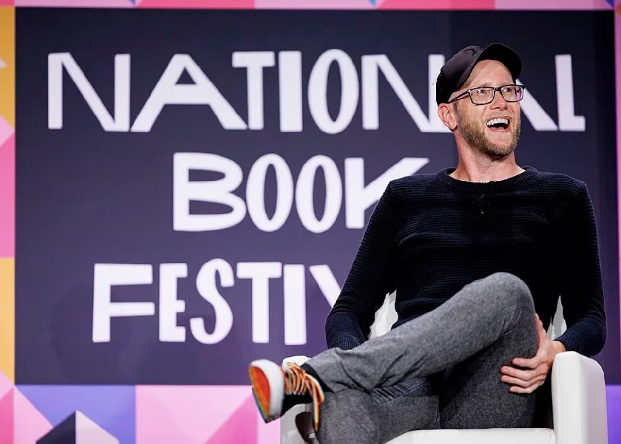 Author TJ Klune sits on stage at the National Book Festival. He is laughing and wearing a black ball cap, long-sleeved black shirt, and jeans. He is white with a close-cut beard and glasses.
