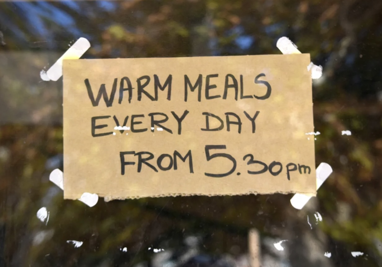 a cardboard sign is taped to a window that reads: Warm Meals Every Day from 5:30 pm