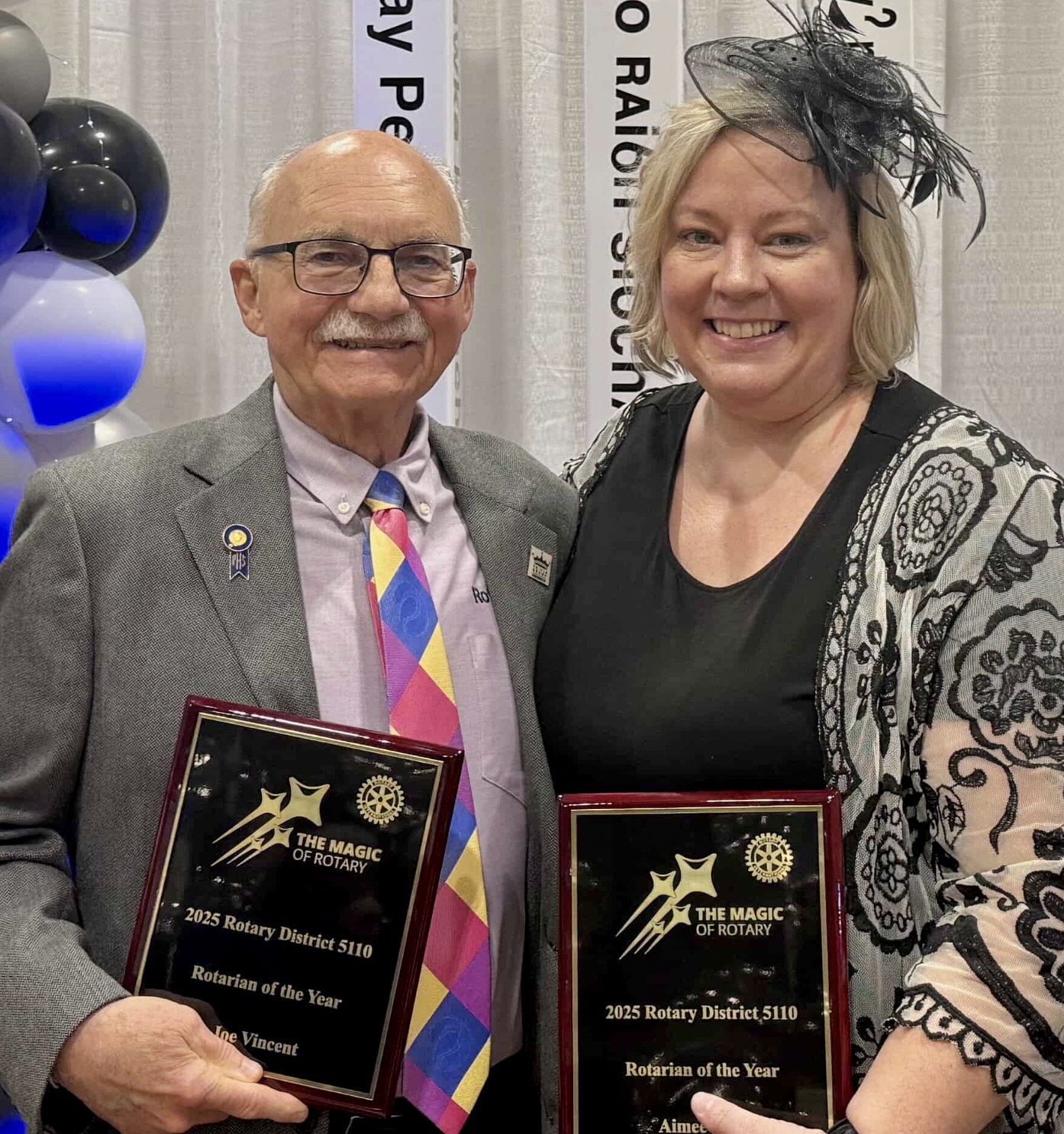 Two Rotarians smile at the camera holding their 2025 Rotarians of the Year awards. She is wearing a black dress and black fascinator with an embroidered shawl. She has chin-length blond hair. He s wearing glasses and a tweed blazer with a bold-colored tie and lavender collared shirt.