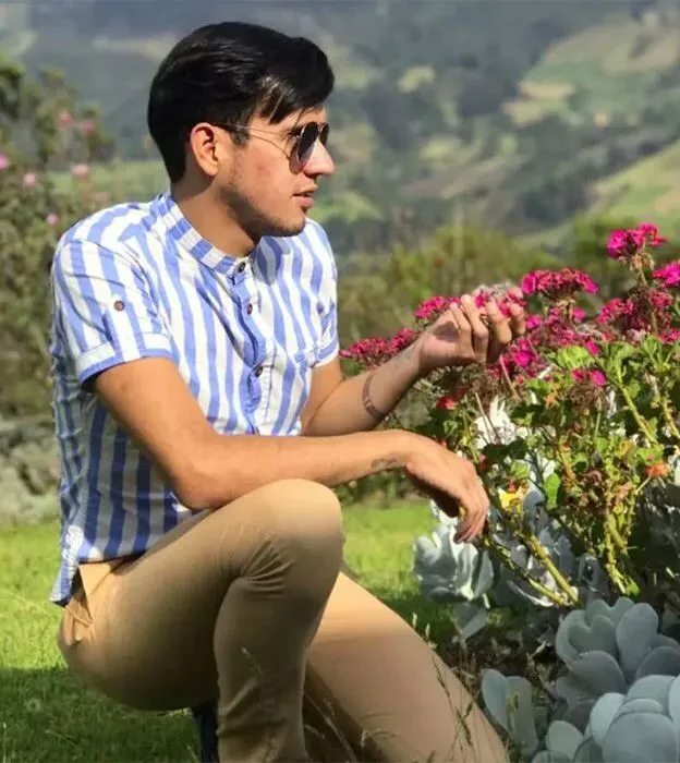 Andry José Hernández Romero is an El Salvadoran kidnap victim of ICE. He is pictured kneeling before some red flowers and cupping some in his hand. The sun is shining and it's grassy behind him. He has short black hair and is wearing sunglasses and a short-sleeved blue and white button-up shirt.