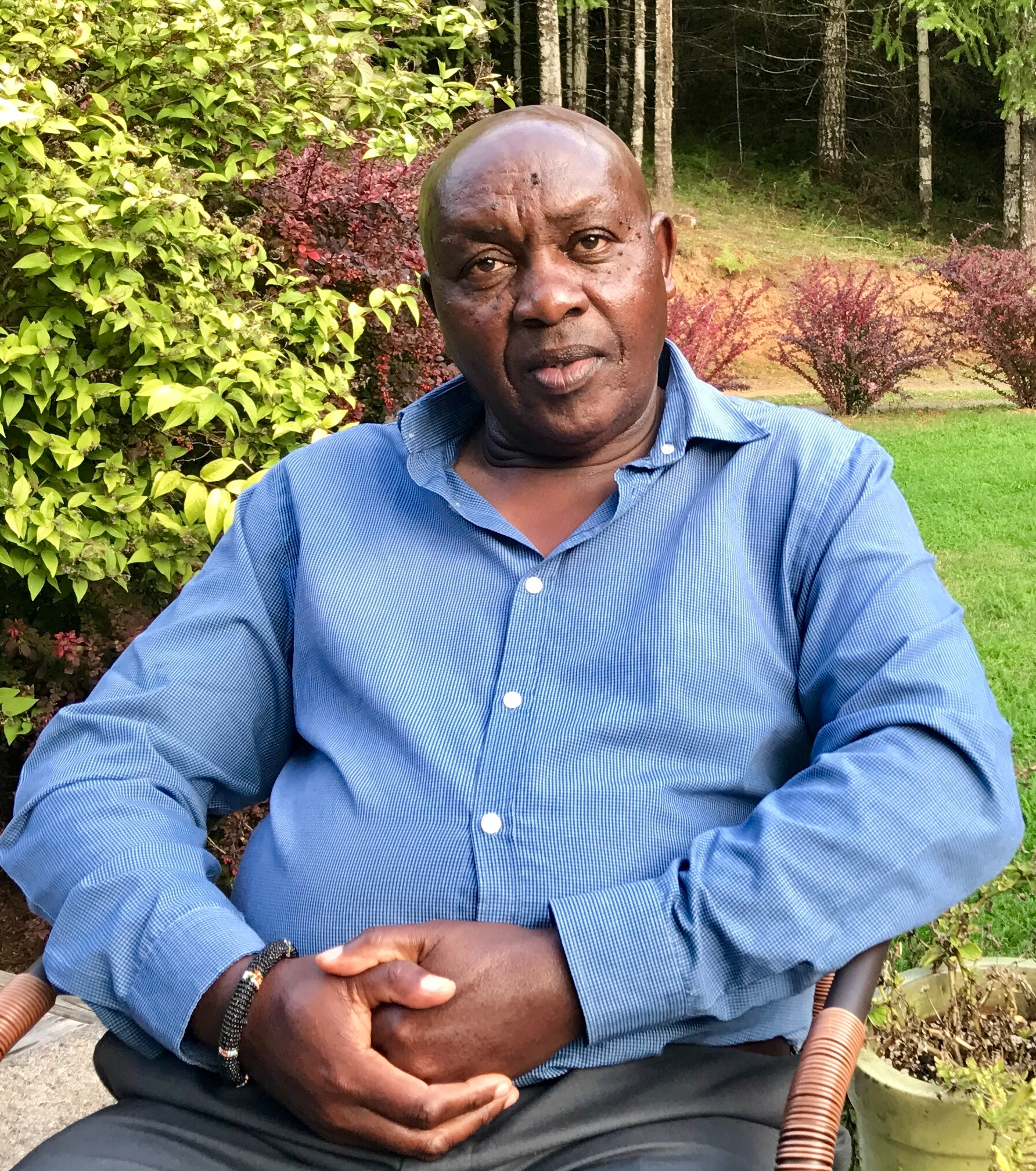 Samuel Muhunyu is wearing a long-sleeved blue button-up shirt. He is sitting outside in Oregon looking directly at the camera with his arms folded in his lap. He is a bald middle-aged Kenyan man.
